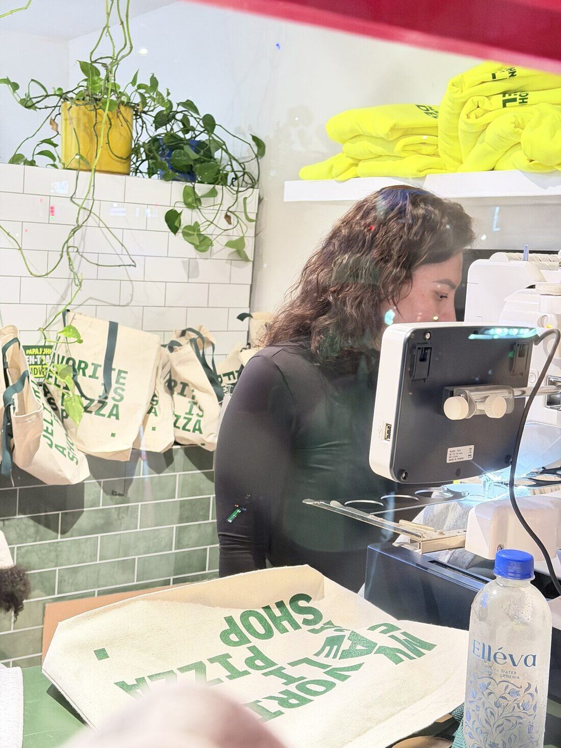A Merch Troop crew member at an embroidery machine stitching a custom tote bag on site, finished totes hanging on the rail behind